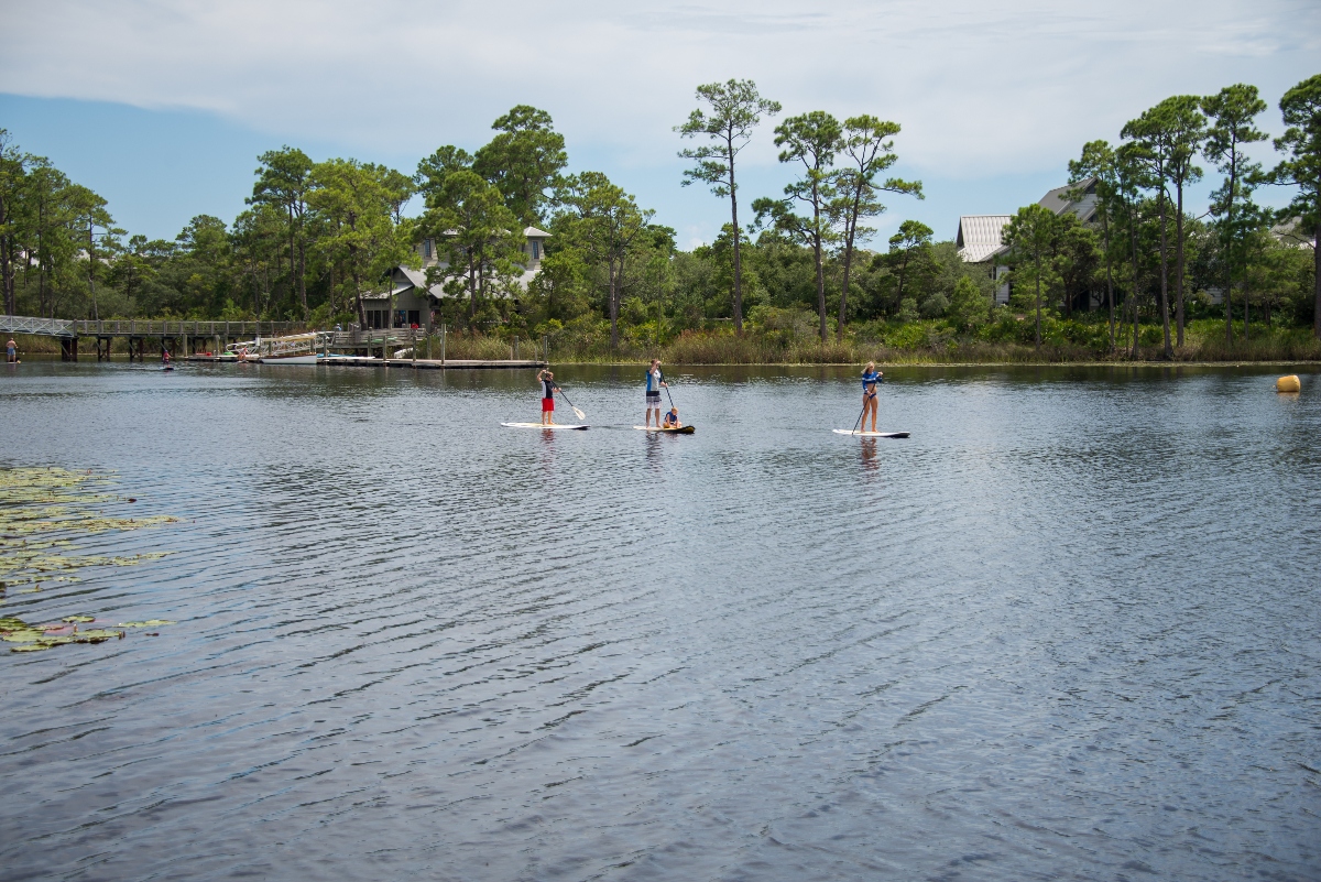 Three people stand on paddleboards in the middle of a calm lake or pond, with a wooden dock visible on the left shore. The scene is surrounded by dense pine and deciduous trees with residential buildings visible in the background under an overcast sky.