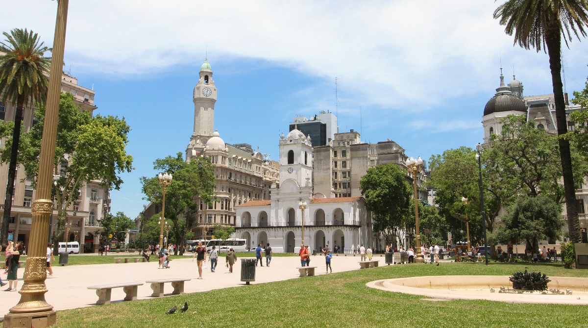 The historic Cabildo building anchors this view of Plaza de Mayo in Buenos Aires, its white colonial facade featuring a row of arches and a central bell tower. The plaza's green lawn and wide pedestrian paths accommodate visitors strolling through the public square, framed by tall palm trees on either side. Behind the Cabildo, the city's European-influenced architecture rises against a partly cloudy sky, including a distinctive clock tower and domed buildings.