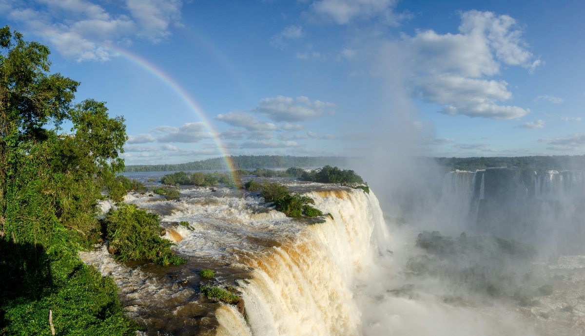 A rainbow arcs across the mist-filled gorge of Iguazu Falls, where multiple cascades of tan-colored water plunge over rocky cliffs bordered by dense subtropical forest. Vegetation-covered islands punctuate the waterfall system, with powerful torrents flowing around and over the basalt formations. Heavy mist rises from the falling water, partially obscuring the far side of the gorge beneath scattered clouds in the blue sky.