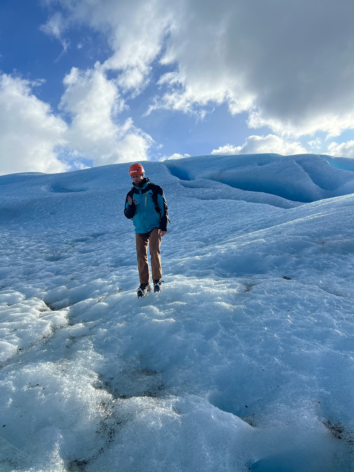 A hiker equipped with crampons stands on the textured blue ice surface of Perito Moreno Glacier, surrounded by the glacier's distinctive rippled formations and deep crevasses. The person wears technical outdoor gear suitable for glacier trekking, positioned against a backdrop of sculpted ice ridges that rise dramatically behind them. White clouds drift across the bright sky above the intensely blue glacial ice, highlighting the scale and otherworldly landscape of this Patagonian ice field.