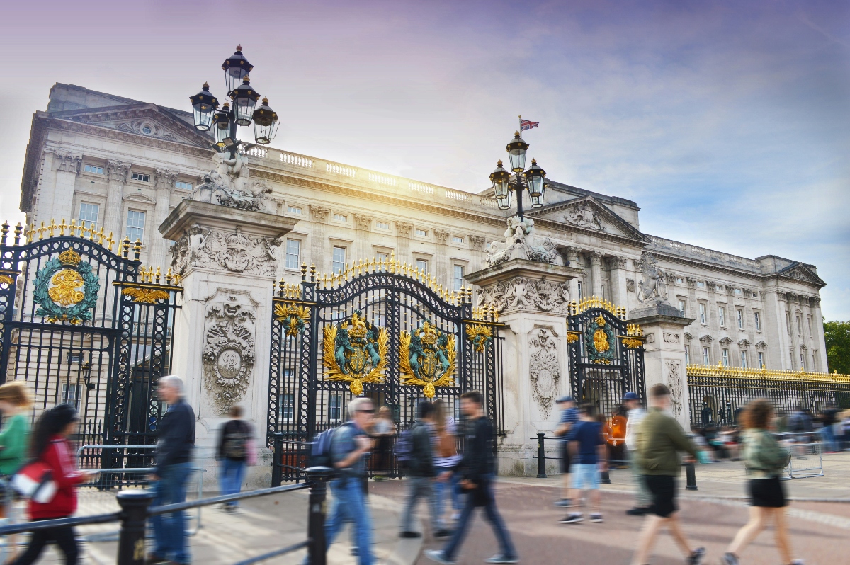 The image shows a grand neoclassical building with light-colored stone facade and classical columns, fronted by ornate black iron gates decorated with gold detailing and royal emblems. Blurred figures of tourists and pedestrians walk past the elaborate entrance gates, which feature intricate metalwork and are flanked by decorative lamp posts under a partly cloudy sky.