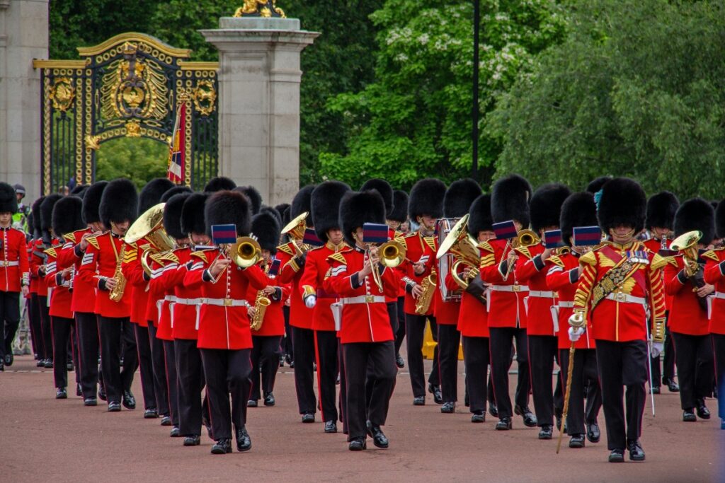 A large military band dressed in red ceremonial tunics and tall black bearskin hats marches in formation while playing brass instruments including trumpets, trombones, and tubas. Behind the musicians, ornate black and gold gates with decorative royal emblems are visible against a backdrop of lush green trees, with the band performing on what appears to be a courtyard or parade ground.