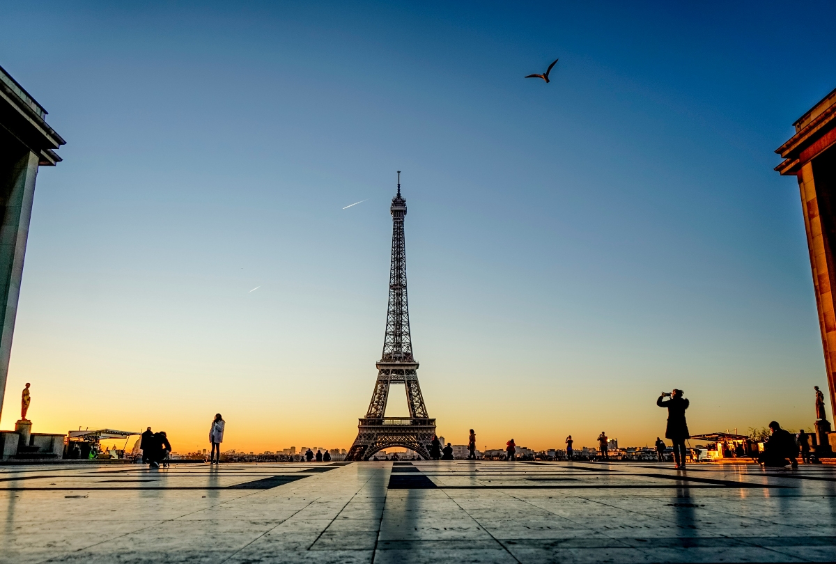 Shot from a low angle across the Trocadéro esplanade, the Eiffel Tower stands centered against a sky graduating from orange at the horizon to deep blue overhead. Scattered visitors appear in silhouette along the paved plaza, one raising a camera, while a bird flies in the upper right of the frame.