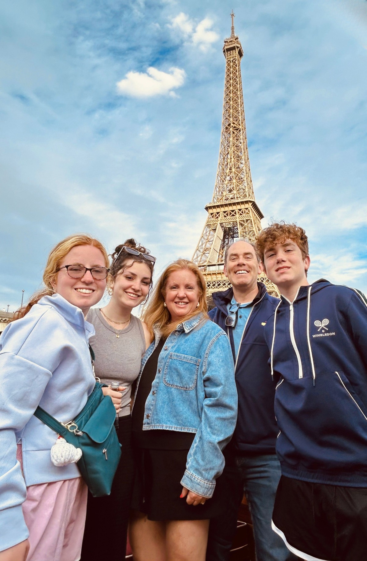 A family of five stands together smiling at the camera, with the Eiffel Tower rising behind them against a blue sky streaked with wispy clouds. Two teenage girls, a woman in a denim jacket, a man in a navy jacket, and a teenage boy in a navy zip-up hoodie are grouped shoulder to shoulder in the lower half of the frame.