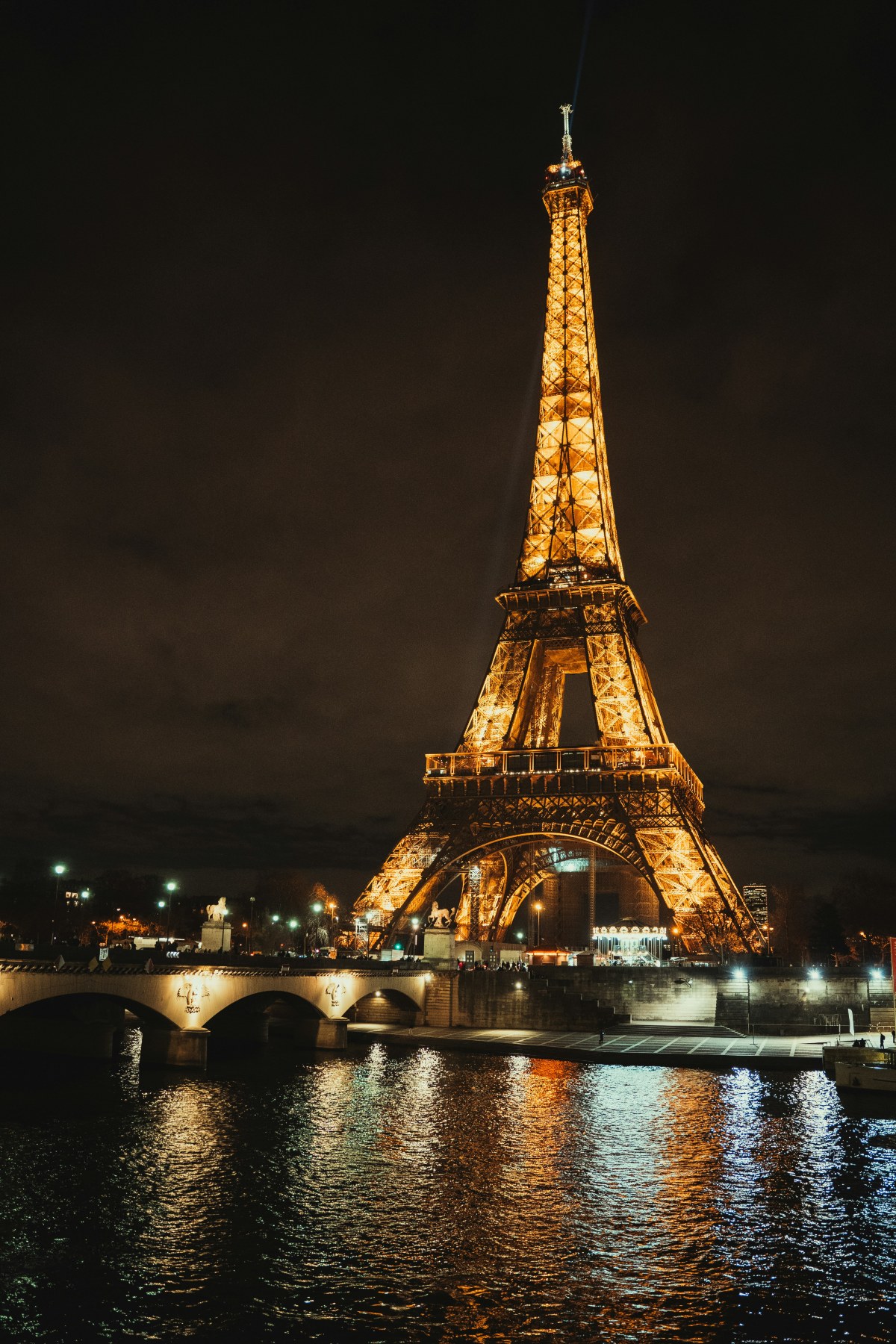 The Eiffel Tower stands illuminated in gold light against a black night sky, with a searchlight beam projecting upward from its summit. In the foreground, the Seine reflects the tower's amber glow in rippling streaks, while a lit stone arch bridge crosses the river at left.