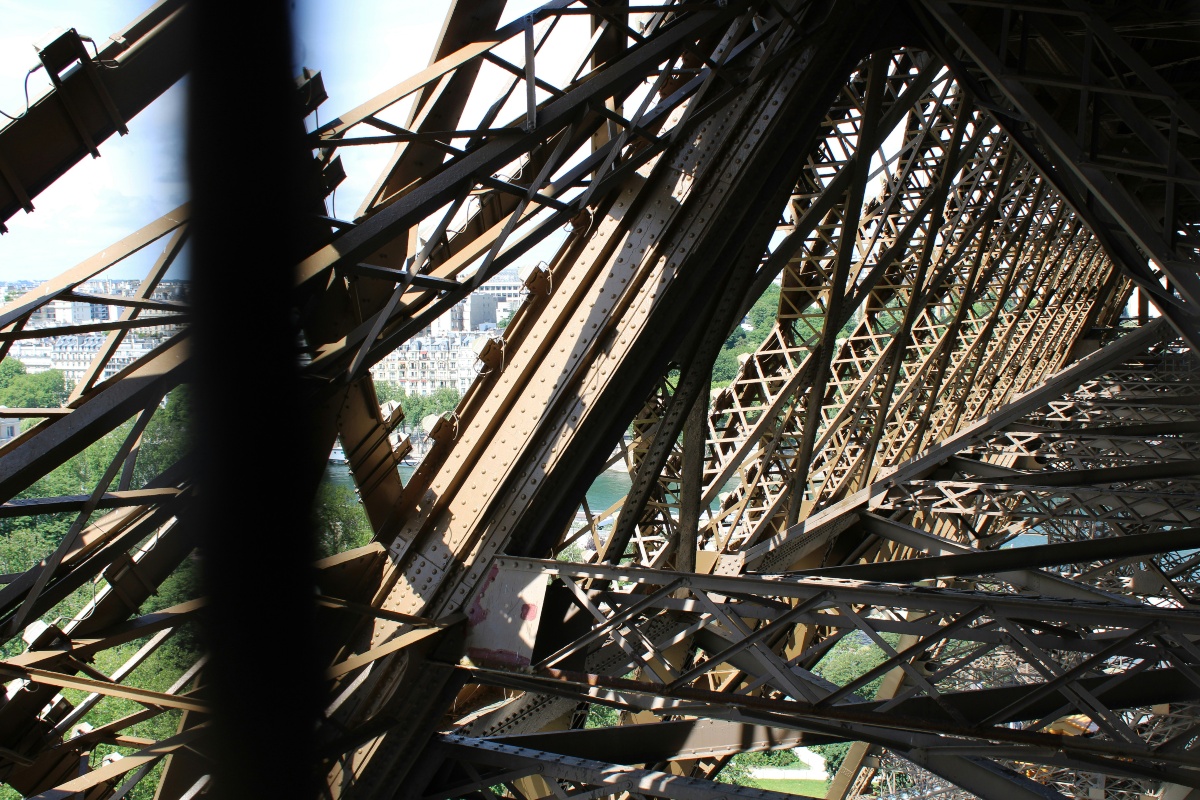 A close view from inside the Eiffel Tower shows a dense lattice of riveted iron beams and cross-bracing angling in multiple directions. Through gaps in the ironwork, the Seine, green treetops, and pale Parisian buildings are visible under a bright sky.