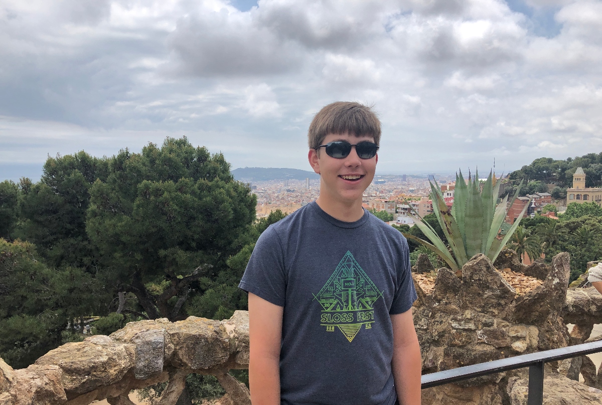 A young person wearing sunglasses and a navy blue "Sloss Fest" t-shirt stands on a stone wall or ledge, smiling at the camera. Behind them is a panoramic view of a city with red-roofed buildings stretching into the distance, framed by lush green trees and vegetation including what appears to be an agave plant.