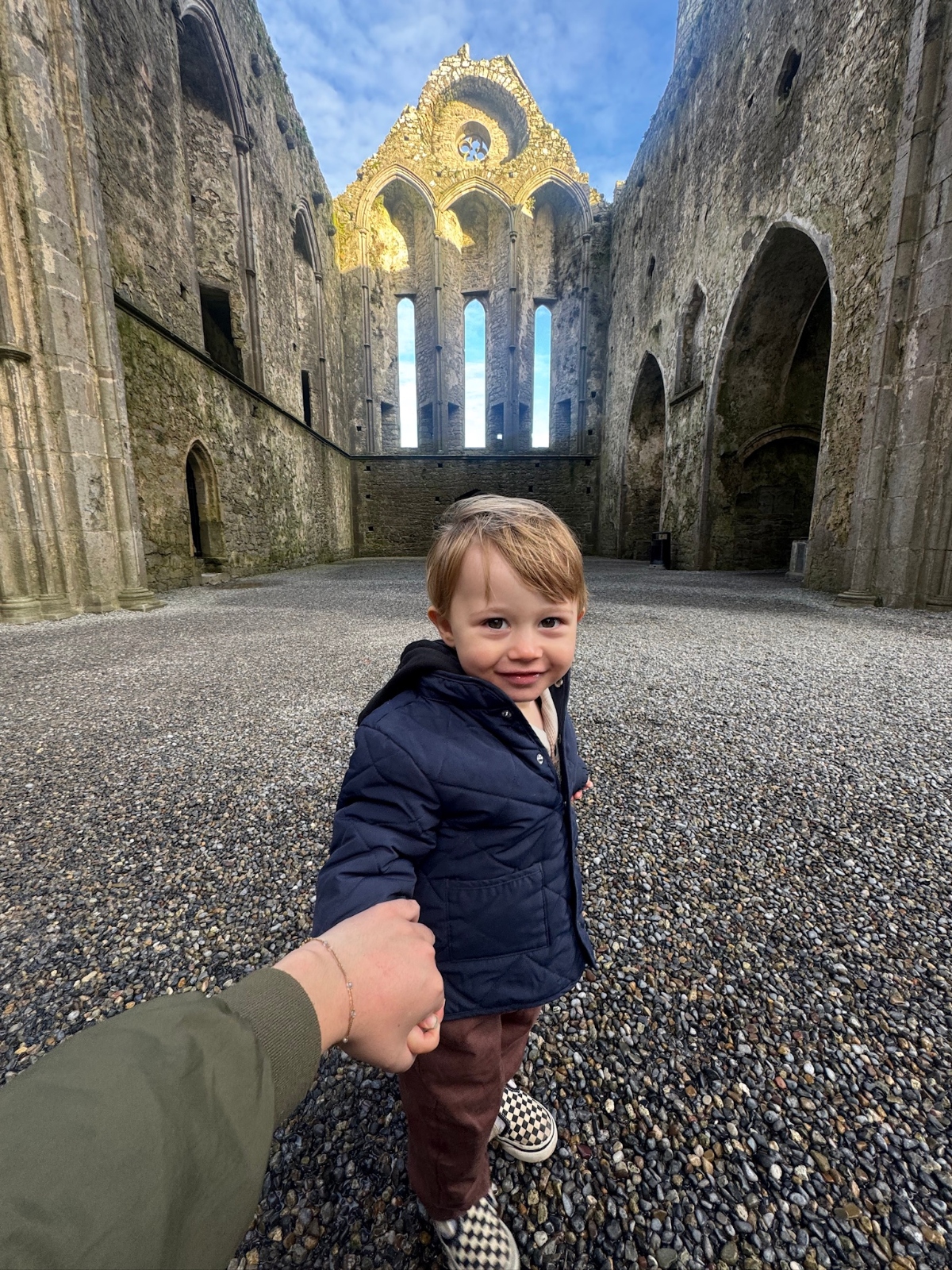 A toddler in a navy jacket and brown pants holds hands with an adult while standing in the interior of a roofless stone abbey or church with tall Gothic arched walls and windows. The medieval structure features weathered gray stone walls with decorative golden stonework at the top, and the ground is covered in gravel beneath an open sky.