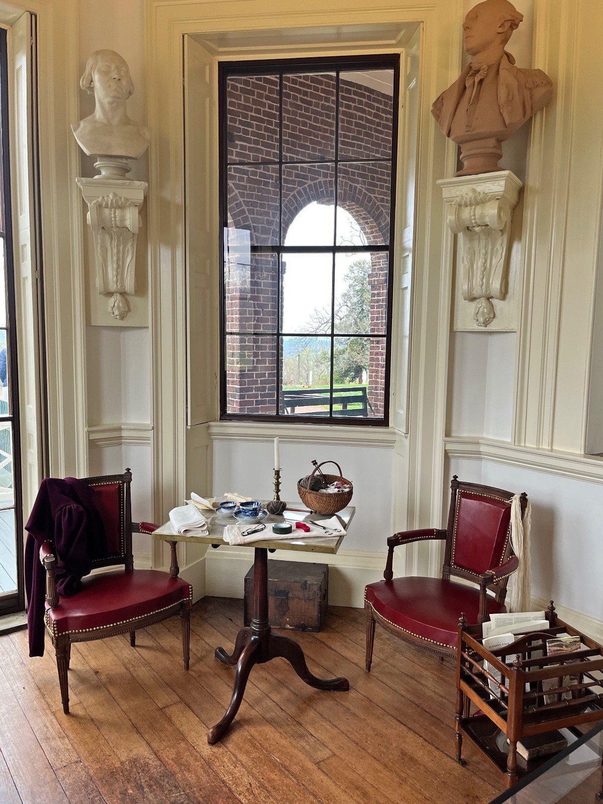 Alt text:A corner sitting room at Thomas Jefferson's Monticello features two red leather armchairs with brass nail trim flanking a small pedestal table set with blue and white porcelain, a candlestick, and a wicker sewing basket. Two white plaster portrait busts are mounted on decorative corbels on either side of a tall multi-pane window overlooking the brick exterior arcade and tree-lined grounds.