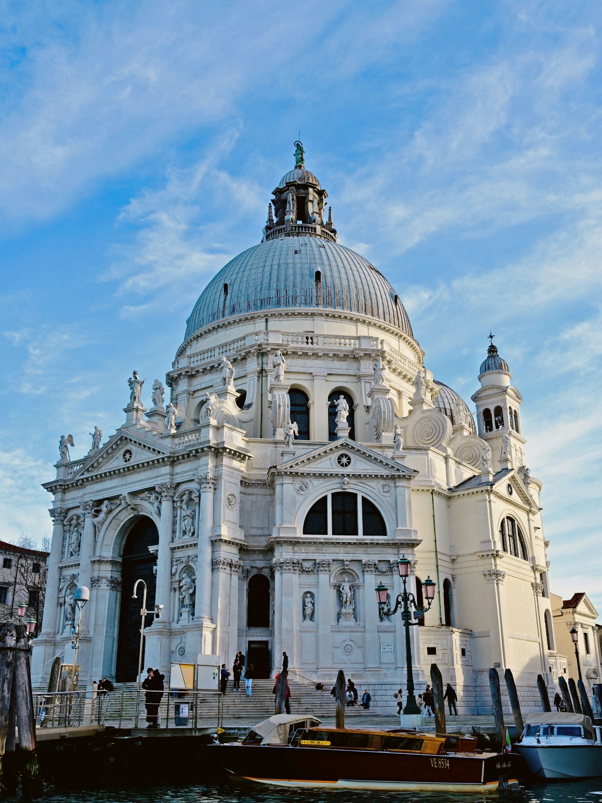 Exterior view of the Basilica di Santa Maria della Salute in Venice, Italy, a 17th-century Baroque church with a large grey dome, ornate white stone facade adorned with classical sculptures, and arched entryways, as seen from the Grand Canal. In the foreground, wooden boats are moored along the waterfront where visitors climb the church's wide stone steps beneath a partly cloudy blue sky.