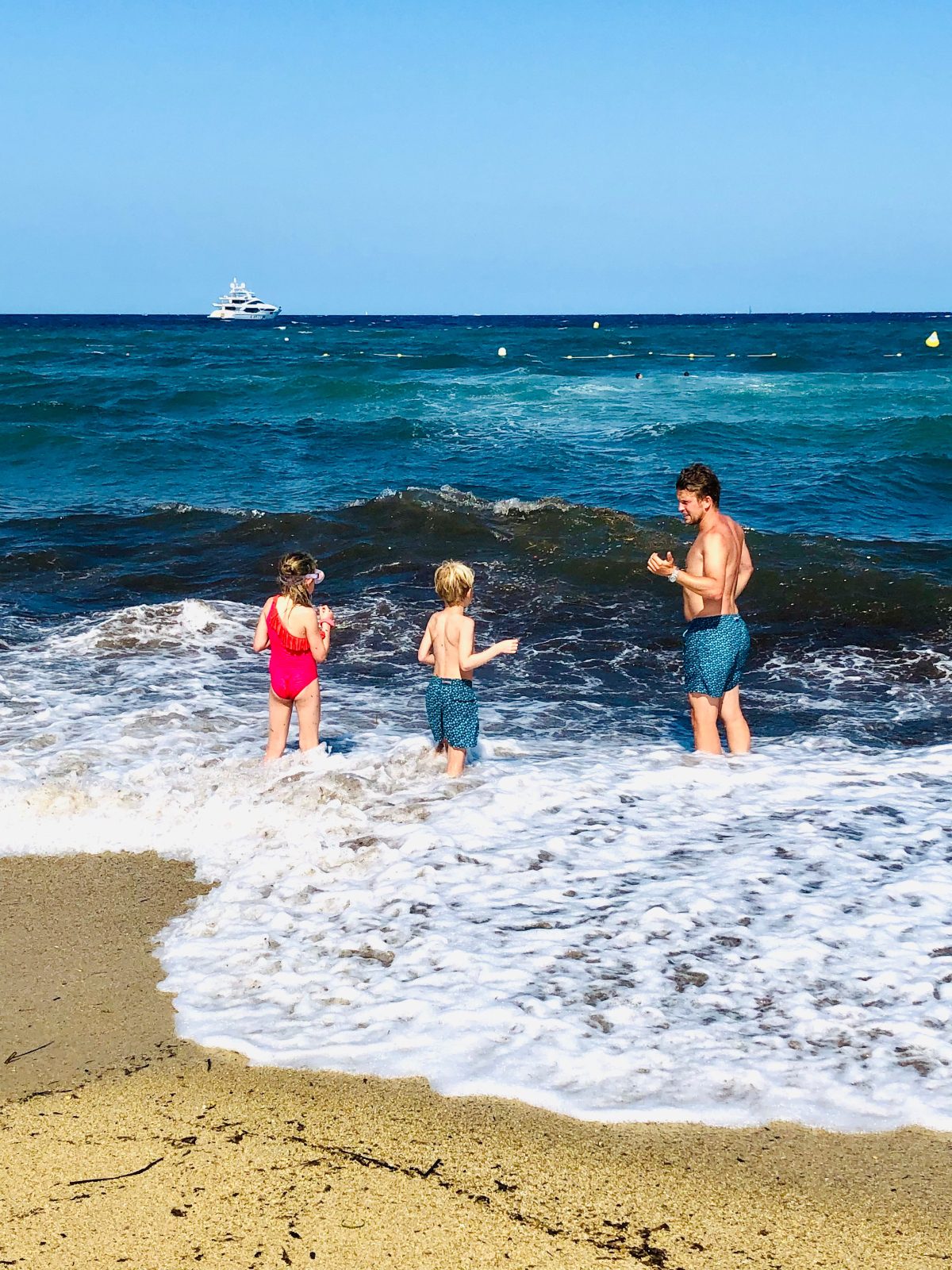 A man wearing a blue print bathing suit stands in shallow surf on a golden beach, accompanied by a young girl in a red bathing suit and a young boy in a blue print bathing suit. A boat is visible in the distanct ocean.