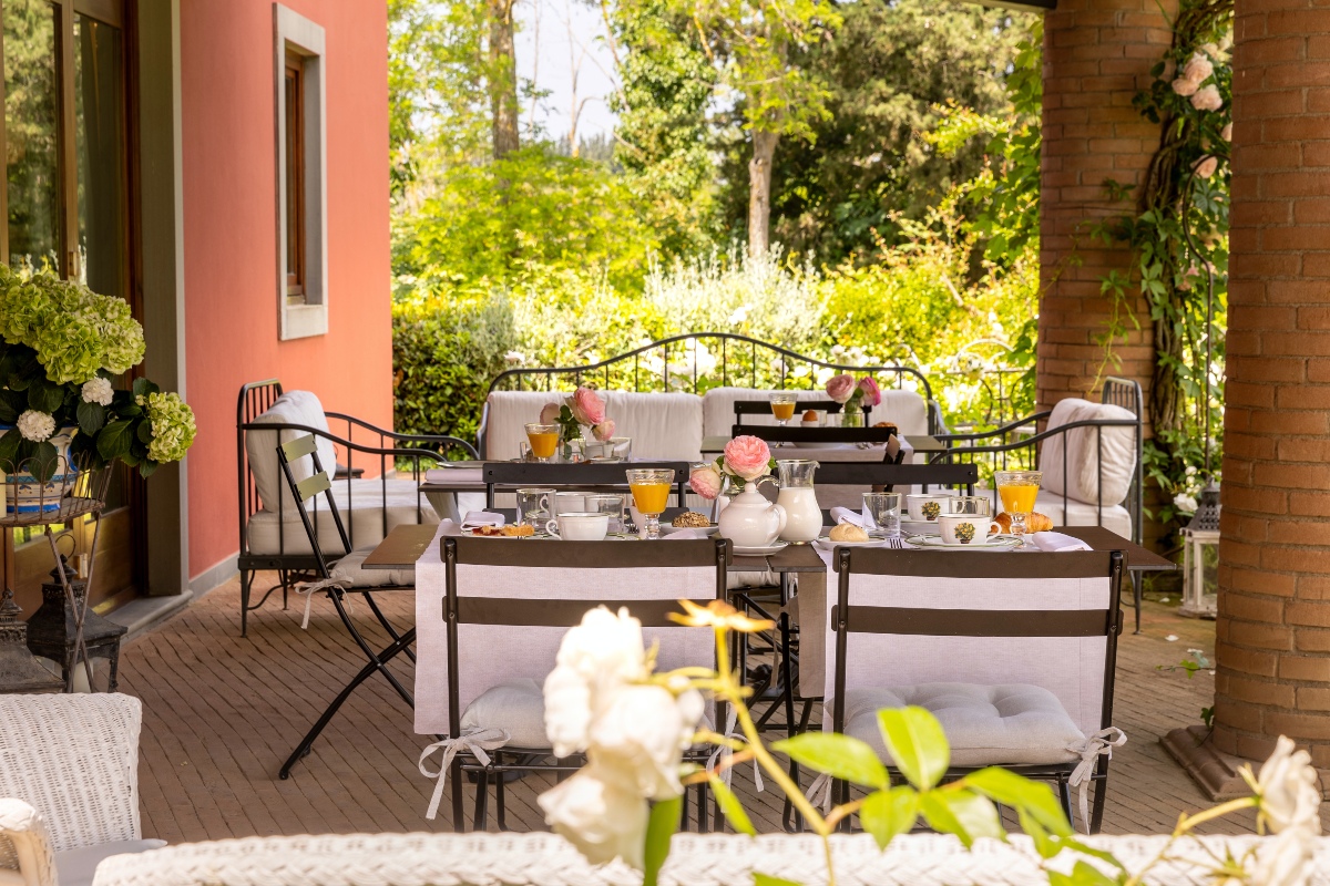 Breakfast is served on the Tuscan farmhouse's outdoor terrace with an iron bistro table, with orange juice, espresso cups, croissants and pink roses in small vases surrounded by brick columns covered with climbing vines. Lush green gardens and terracotta pink villa facades form the backdrop, while white hydrangeas and garden flowers fill the foreground.
