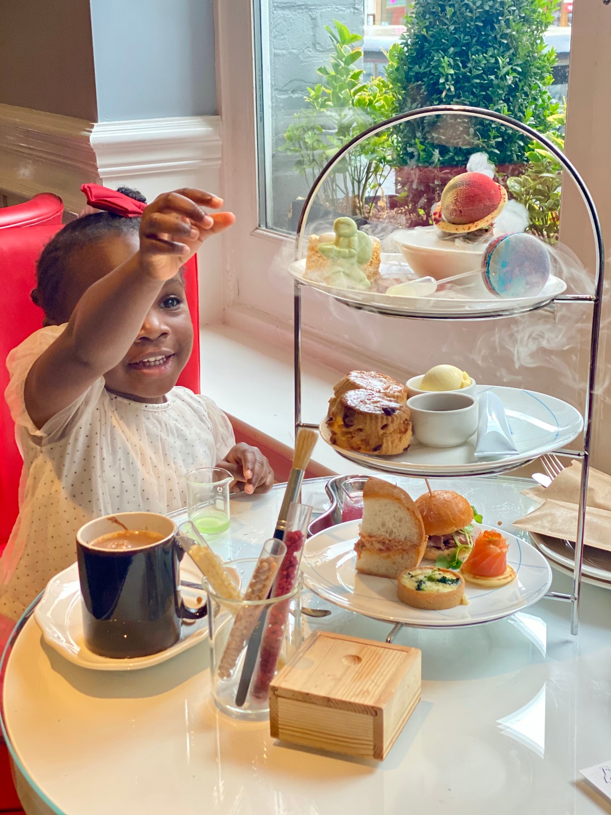 A young child in a white dotted blouse and red hair bow sits at a small round table, smiling and reaching upward with one hand. In front of her is a three-tiered afternoon tea stand holding finger sandwiches and a mini quiche on the bottom plate, a scone with clotted cream and jam in the middle, and colorful pastries on top, with a mug, a glass of green liquid, test tubes of sprinkles, and a small wooden box arranged on the table beside it.