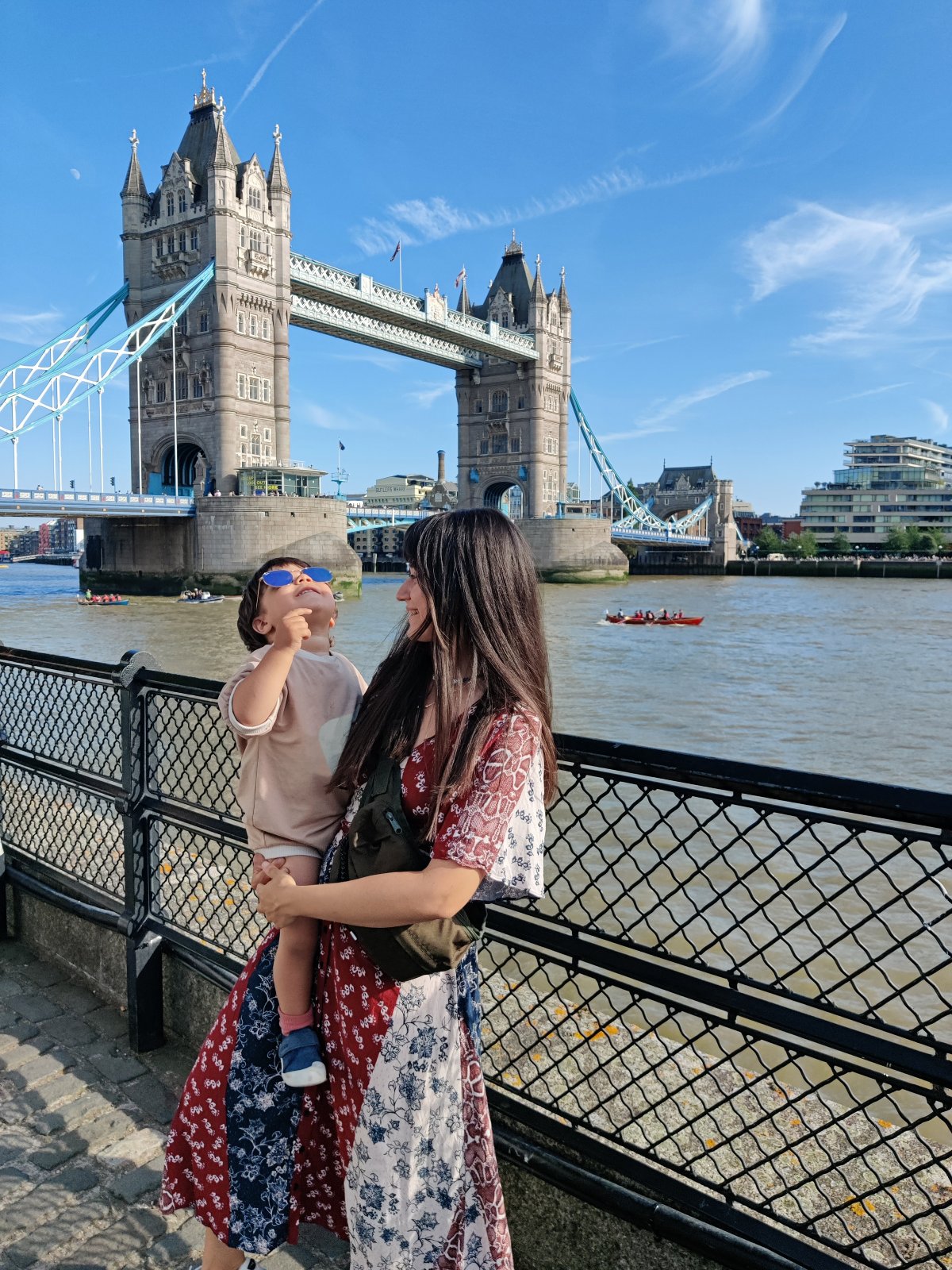 A woman in a red and blue floral dress stands at a railed walkway along the River Thames, holding a toddler in blue sunglasses and a beige outfit who tilts his head back to look upward. Tower Bridge rises behind them under a clear blue sky, with rowing boats crossing the brown water and riverside buildings lining the far bank.