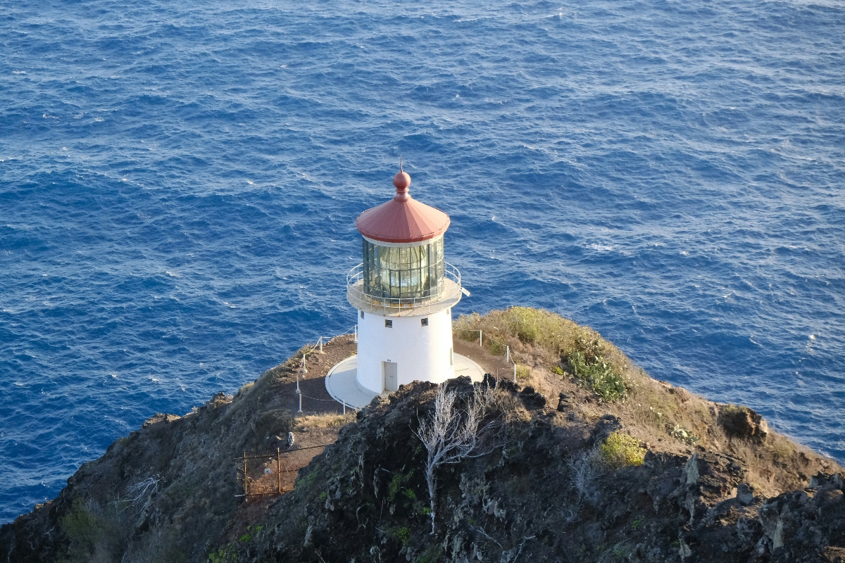 This aerial view shows a white cylindrical lighthouse with a red dome roof and glass lantern room positioned on a small rocky outcrop surrounded by deep blue ocean waters. The lighthouse stands on rugged terrain with sparse vegetation and visible pathways, completely isolated from any mainland and encircled by the open sea.
