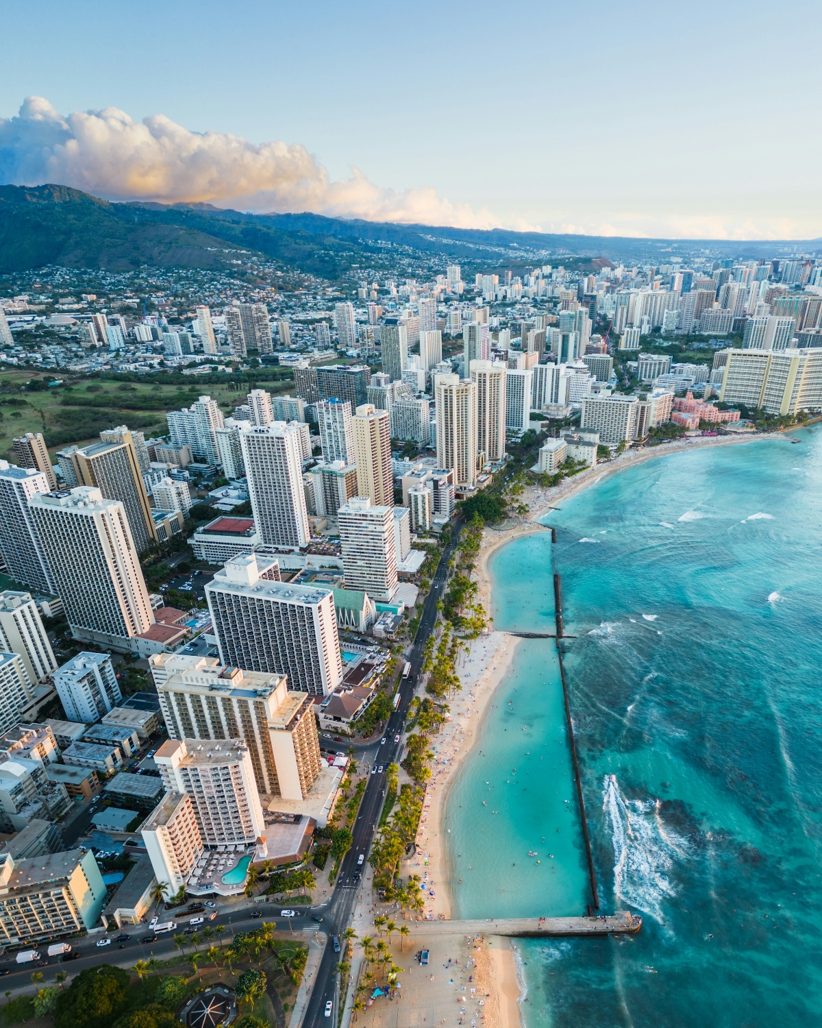 This aerial photograph captures a densely developed coastal city with numerous high-rise buildings and hotels extending along a pristine sandy beach, while a pier or breakwater structure extends into the crystal-clear turquoise ocean waters. The urban landscape is framed by lush green mountains in the background and scattered white clouds in a blue sky, with palm trees lining the beachfront road that separates the buildings from the shore.