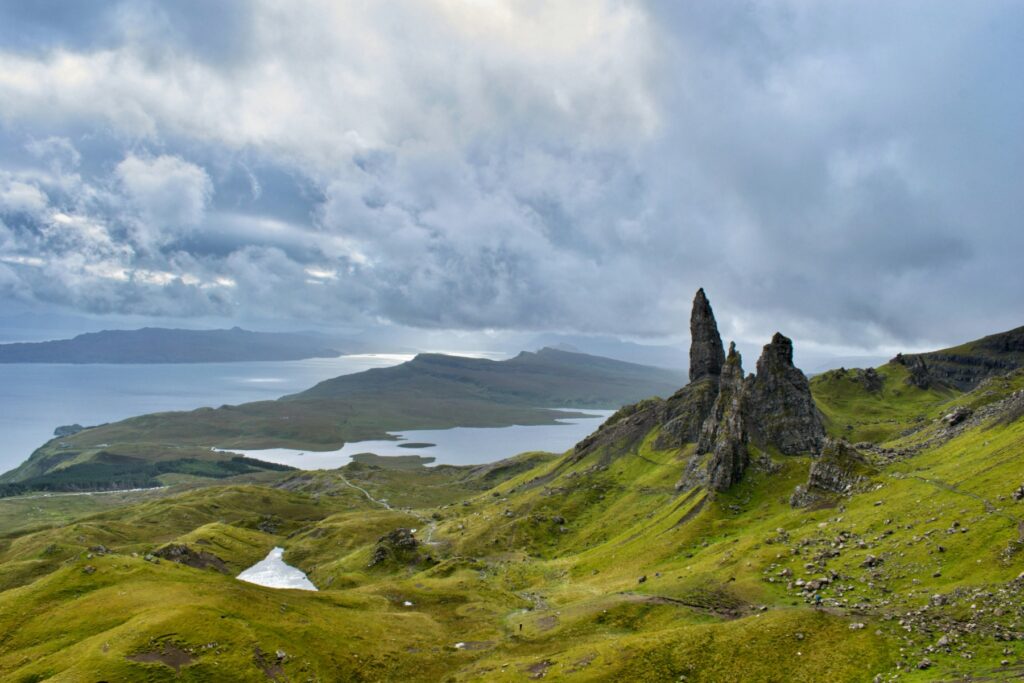 Tall, jagged basalt pinnacles rise from a steep green hillside at the Old Man of Storr on the Isle of Skye, with rolling emerald moorland and lochs stretching toward the coast below. Heavy storm clouds fill most of the sky above, while the Sound of Raasay and a distant island headland are visible across the water in the background.