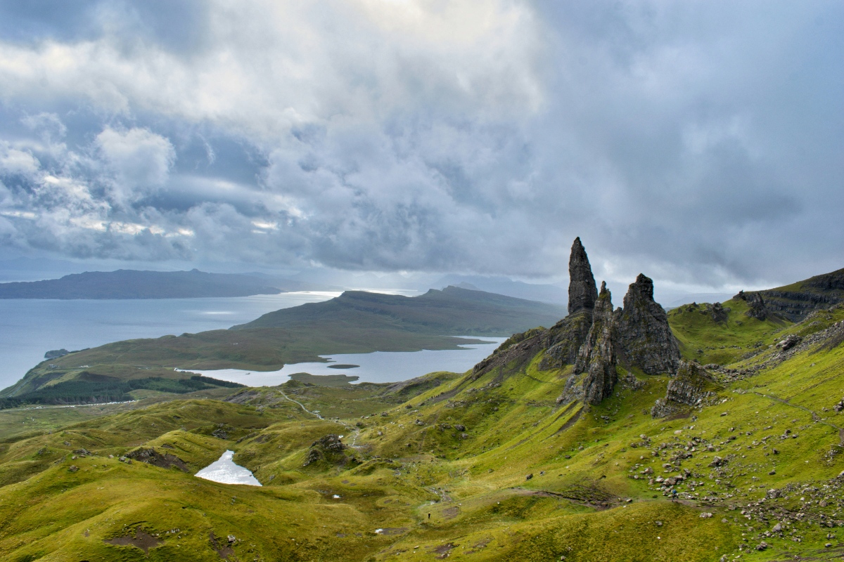 Tall, jagged basalt pinnacles rise from a steep green hillside at the Old Man of Storr on the Isle of Skye, with rolling emerald moorland and lochs stretching toward the coast below. Heavy storm clouds fill most of the sky above, while the Sound of Raasay and a distant island headland are visible across the water in the background. 