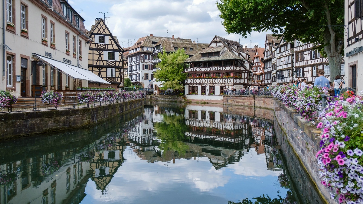 Half-timbered buildings in black and white line a calm canal in Strasbourg's Petite France district, their facades and the partly cloudy sky reflected clearly in the still water below. Pink and purple petunias cascade from the stone bridge railing in the foreground, while pedestrians stroll along the quay on the right.