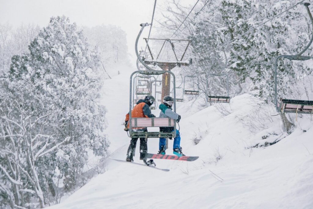 Two riders — one on skis, one on a snowboard — sit on a chairlift as heavy snow falls around them, surrounded by densely packed, snow-covered trees. The slope below and the empty chairs stretching into the misty background suggest an active ski day in low-visibility conditions.