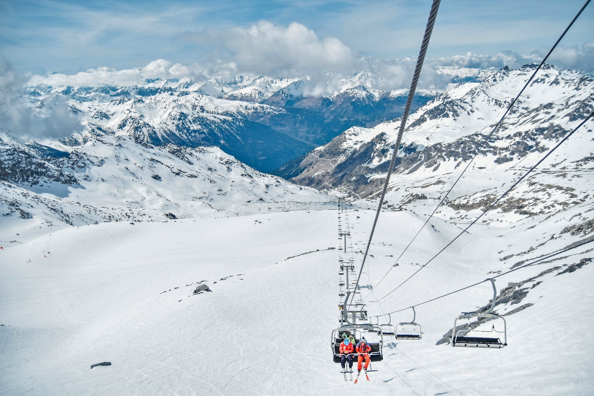 A chairlift carries skiers in brightly colored gear up a high-altitude mountain slope, with the lift cables and multiple chairs visible stretching into the distance. Below lies a vast snow-covered bowl or glacier surrounded by dramatic Alpine peaks under a blue sky with white clouds. The barren, treeless terrain indicates the extreme elevation of this ski area, with mountain ranges visible across multiple horizons.