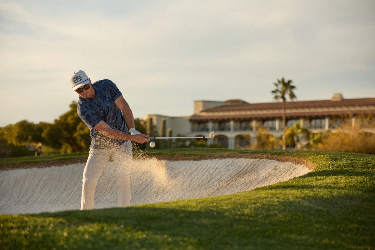 A golfer wearing a blue patterned shirt, white pants, baseball cap, and sunglasses hits a shot from a sand bunker, creating a spray of sand as the ball is struck. The golf course features green grass surrounding the bunker, with resort buildings and palm trees visible in the background under an overcast sky in late afternoon lighting.