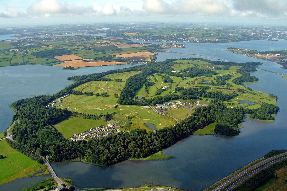 An aerial view shows Fota Island surrounded by water, featuring a mixture of green open spaces, wooded areas, and several buildings including a golf course and hotel complex. The island is connected by a bridge to the mainland, with agricultural fields and residential developments visible across the surrounding landscape under a cloudy sky.