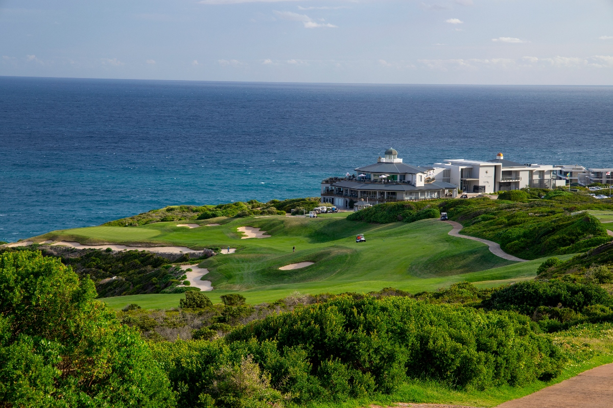 A coastal golf course features multiple green fairways with sand bunkers positioned between dense vegetation and the ocean, with golf carts visible on the course. A modern white clubhouse building and lighthouse structure are situated on the cliffside overlooking the blue water, while tropical vegetation covers the rolling terrain surrounding the course.