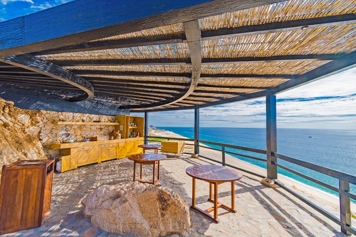 An outdoor terrace features a reed-covered pergola overhead, yellow kitchen cabinets built into a stone wall, and wooden round tables with woven tops positioned on a stone floor. The space overlooks the ocean through glass railings, with a large natural rock formation incorporated into the seating area and blue water visible extending to the horizon.