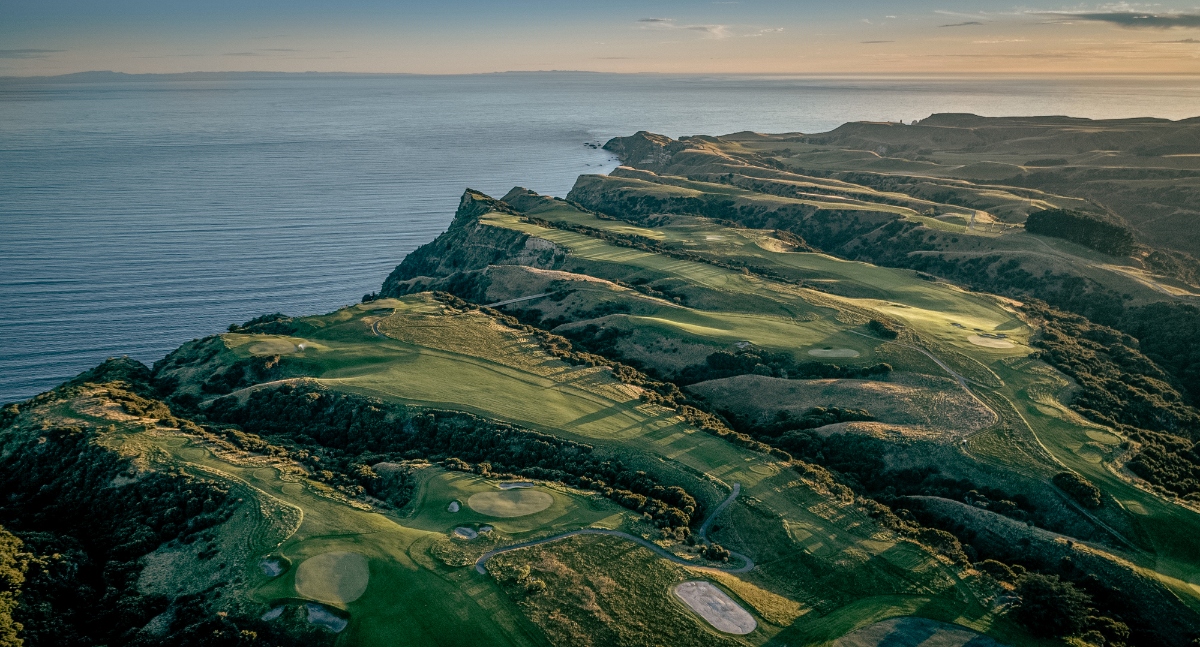 An aerial view shows a golf course built along dramatic coastal cliffs, with multiple holes featuring green fairways, sand bunkers, and water hazards carved into the rolling hillside terrain. The course extends along the clifftops above the ocean, with steep drops to the water below and undulating landscape stretching inland under an expansive sky.