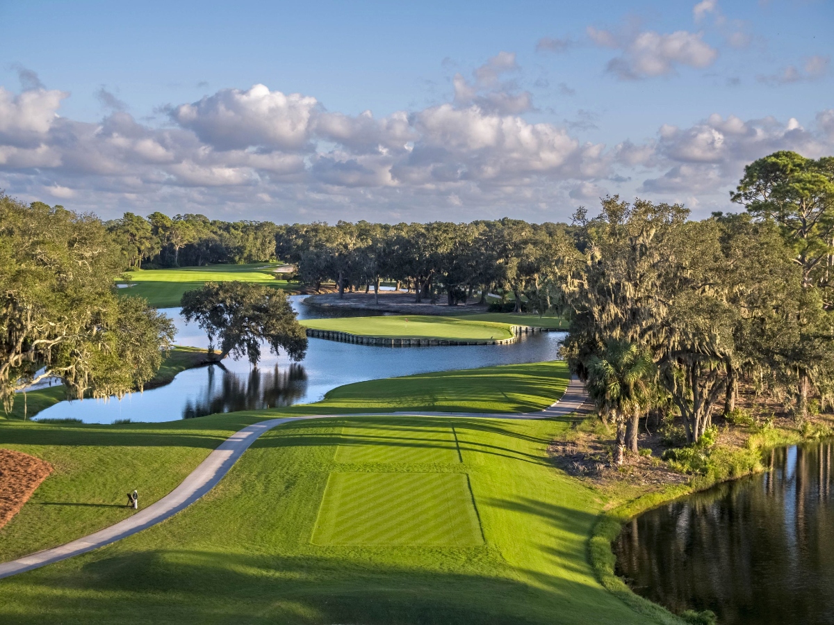 An aerial view shows a golf course with multiple green fairways separated by water features, including ponds and waterways that wind throughout the layout. The course is surrounded by dense trees draped with Spanish moss, palm trees, and other vegetation, with cart paths connecting the various holes under a partly cloudy sky.