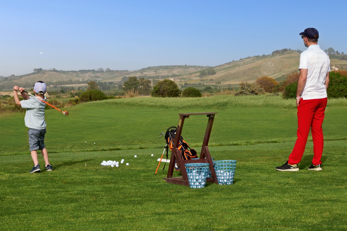 A young golfer in a white shirt and gray shorts practices his swing on a driving range while an adult in red pants and a white shirt watches nearby. The practice area features a wooden ball dispenser with blue baskets, scattered golf balls on the grass, and a golf bag, set against a backdrop of rolling hills under a clear blue sky.