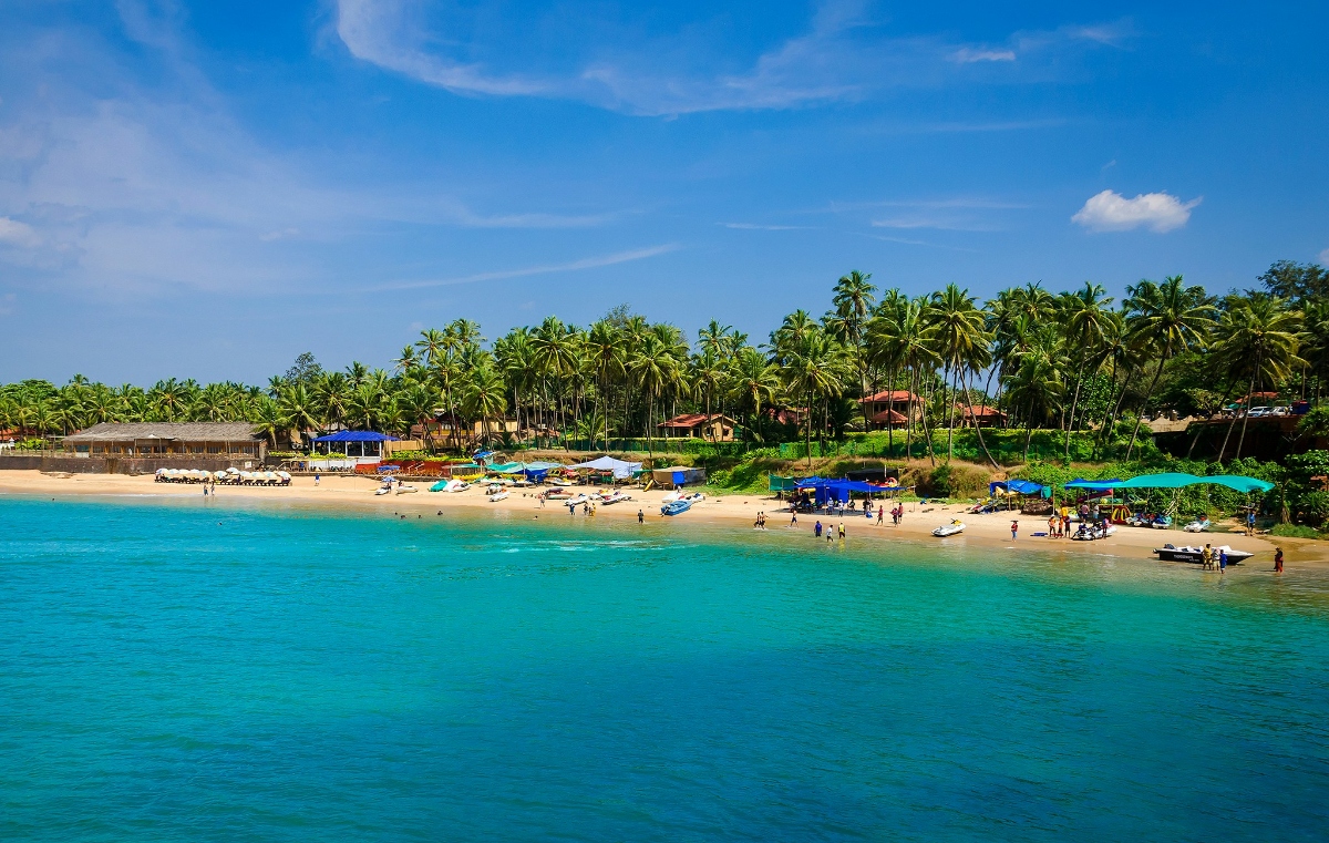 This image shows a curved sandy beach with turquoise ocean water, where numerous people are gathered under colorful beach umbrellas and near various watercraft along the shoreline. The beach is backed by densely forested hills covered with palm trees and tropical vegetation, with several buildings and structures visible among the greenery. The scene is set under a bright blue sky with scattered white clouds.