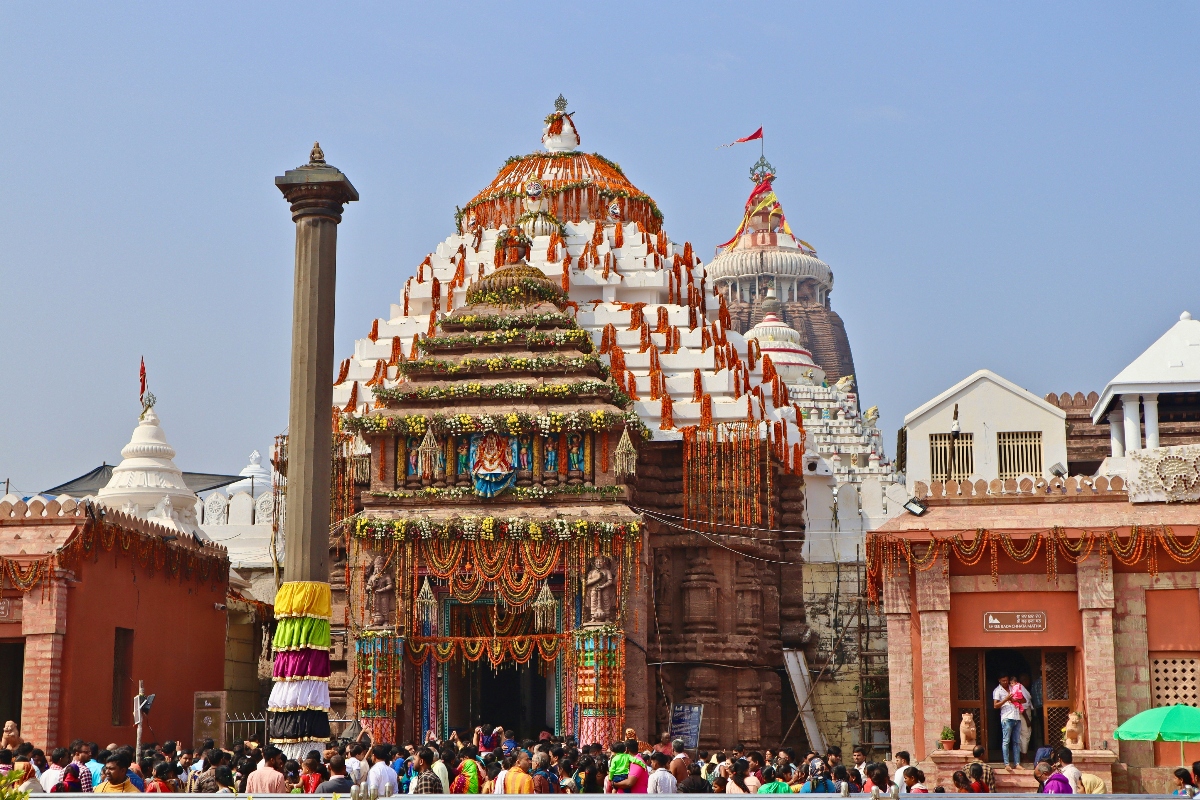 This image shows a large, elaborately decorated multi-tiered wooden structure adorned with colorful garlands, flowers, and religious imagery, surrounded by crowds of people in a temple complex. The structure appears to be a ceremonial chariot or tower with ornate decorations and what look like carved or painted figures displayed on its various levels. The setting includes multiple temple buildings with different architectural styles, including white-domed structures and reddish sandstone buildings, under a clear blue sky.