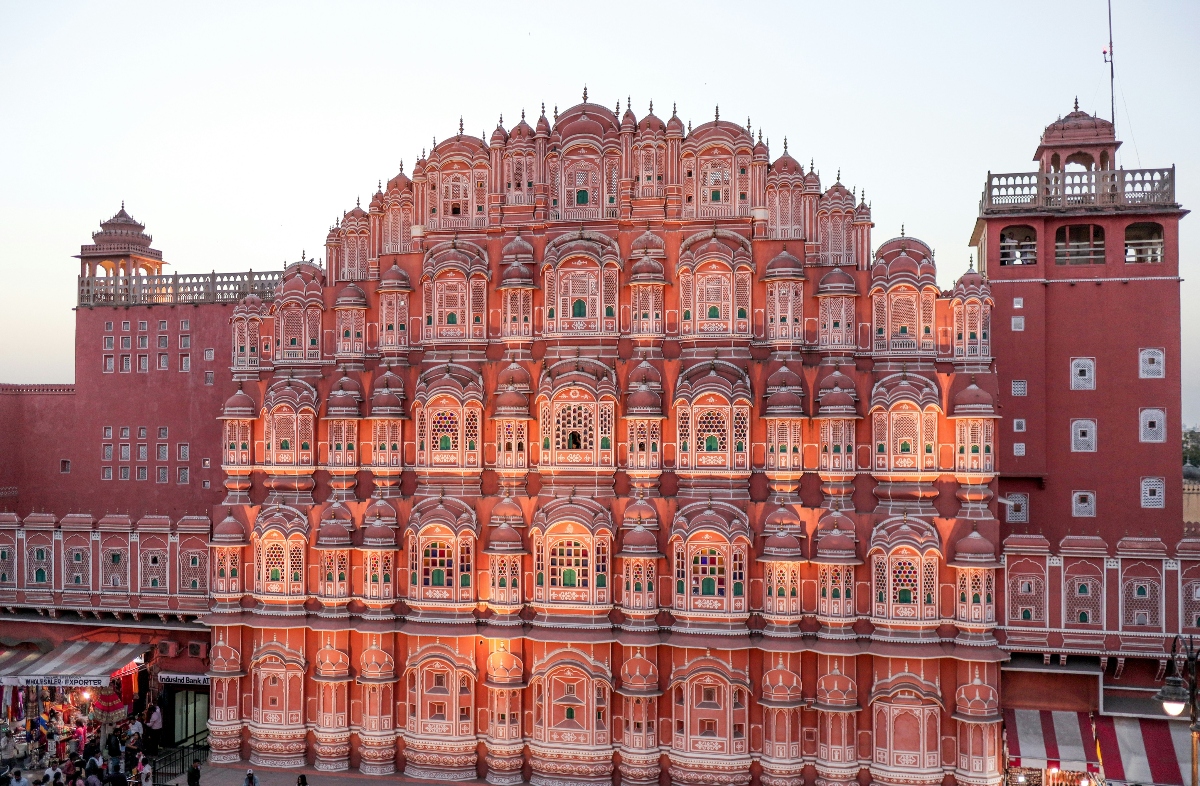 This image shows a large pink sandstone building featuring an ornate facade with hundreds of small arched windows and balconies arranged in multiple tiers, creating a distinctive honeycomb-like pattern. The structure displays characteristic Rajasthani architectural elements including decorative stonework, carved details, and small domed pavilions, with flanking towers visible on either side. The building appears to be illuminated by warm lighting that emphasizes the reddish-pink color of the sandstone construction.