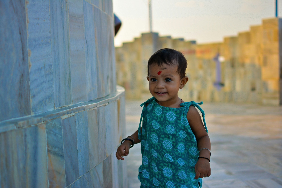 A young child wearing a turquoise patterned sleeveless dress stands beside a weathered blue wall, with a red mark visible on their forehead. The child has dark hair and is wearing what appears to be bracelets or bangles on their wrists. The background shows blurred urban buildings bathed in warm, golden lighting.