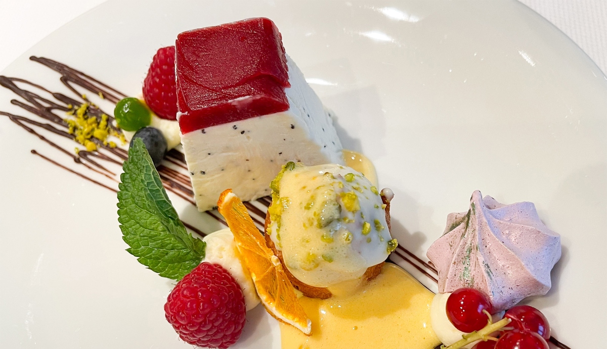 A beautifully plated dessert includes a lavender meringue, a scoop of ice cream dotted with pistachios, and a creamy white cube topped with a layer of magenta fruit spread, on a bed of creme anglaise and chocolate drizzle garnished with a mint leaf.