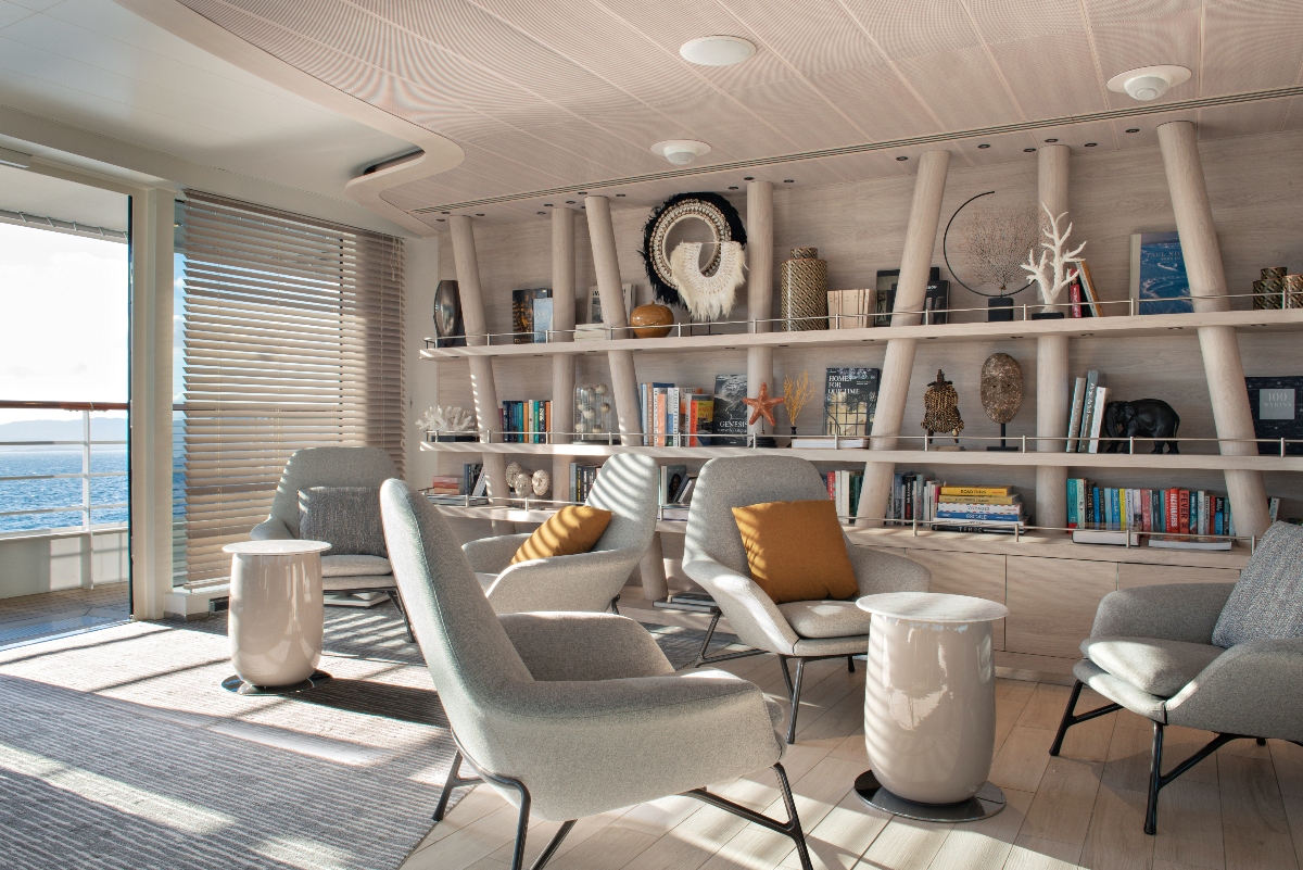 A modern cruise ship library and lounge area featuring contemporary gray upholstered chairs arranged around white cylindrical side tables. The space has floor-to-ceiling built-in white shelving filled with books, decorative artifacts, and collectibles spanning multiple levels. Large windows with venetian blinds offer panoramic ocean views, with natural sunlight streaming across the light wood flooring.
