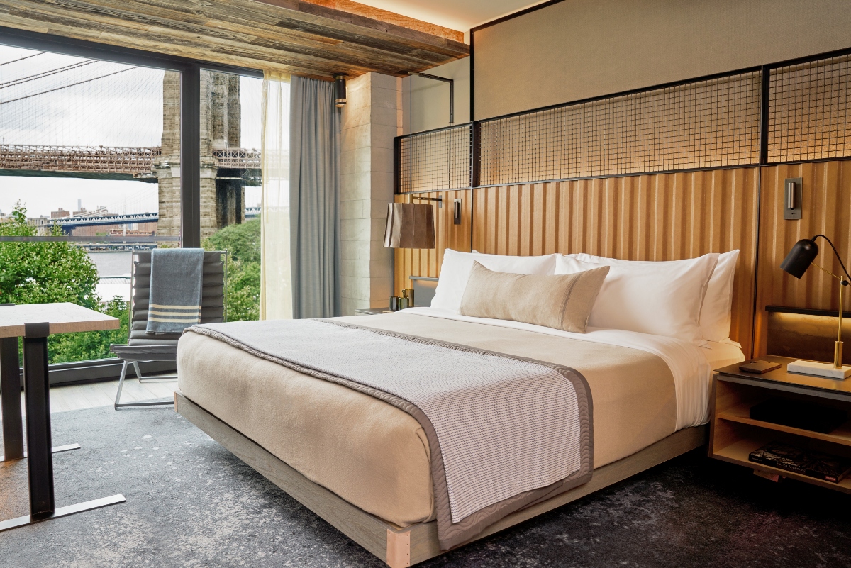 This contemporary hotel room features a king-size bed with cream-colored linens and a distinctive wooden headboard with vertical slats, positioned beneath exposed wooden ceiling beams and a mesh panel detail. Large floor-to-ceiling windows offer views of a historic bridge and waterfront, while the room includes modern furnishings, adjustable bedside lighting, and access to an outdoor terrace or balcony.