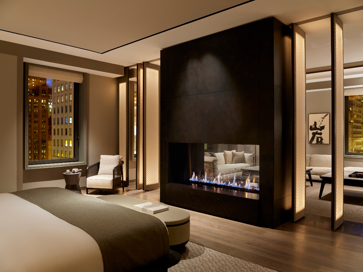 This modern hotel room features a large bed positioned in front of a dramatic black stone fireplace wall with an active linear gas fireplace, while floor-to-ceiling windows reveal illuminated New York City buildings at night. The space includes contemporary furnishings such as a curved upholstered bench at the foot of the bed, a white armchair, and illuminated decorative panels that frame the room's architectural elements.