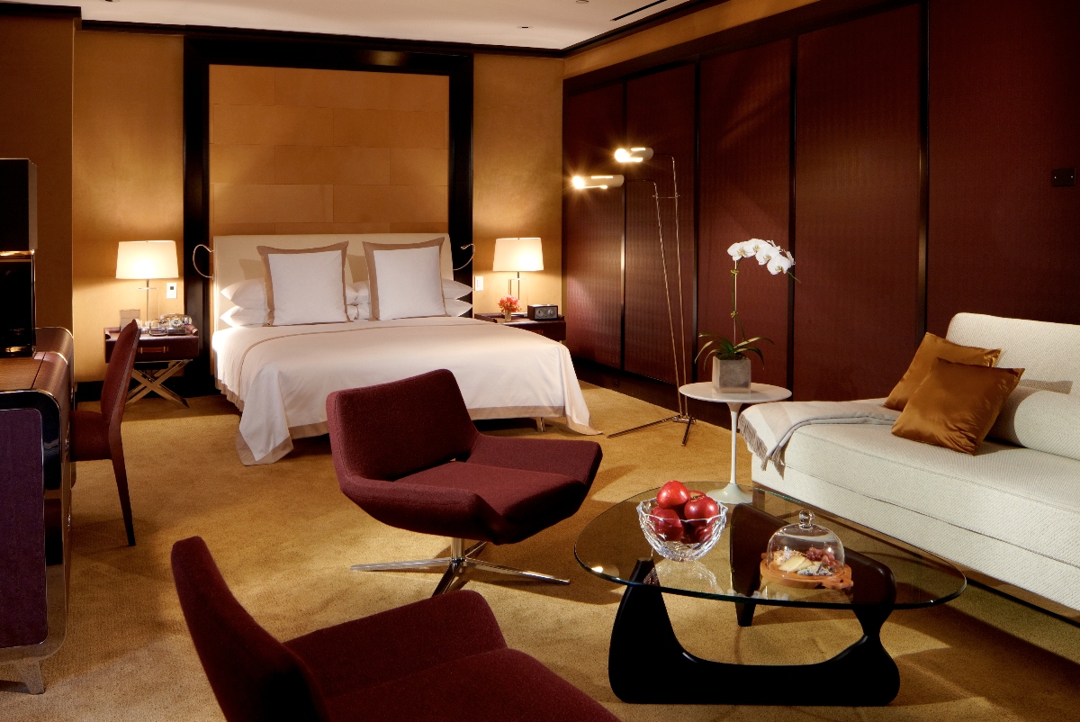 This luxurious hotel suite features a contemporary design with warm gold walls, dark wood paneling, and a bed with white linens set against a tan upholstered headboard framed in dark wood. The spacious room includes a modern seating area with a cream-colored sofa, burgundy swivel chair, and sculptural glass coffee table, all arranged on neutral carpeting with sophisticated lighting throughout.