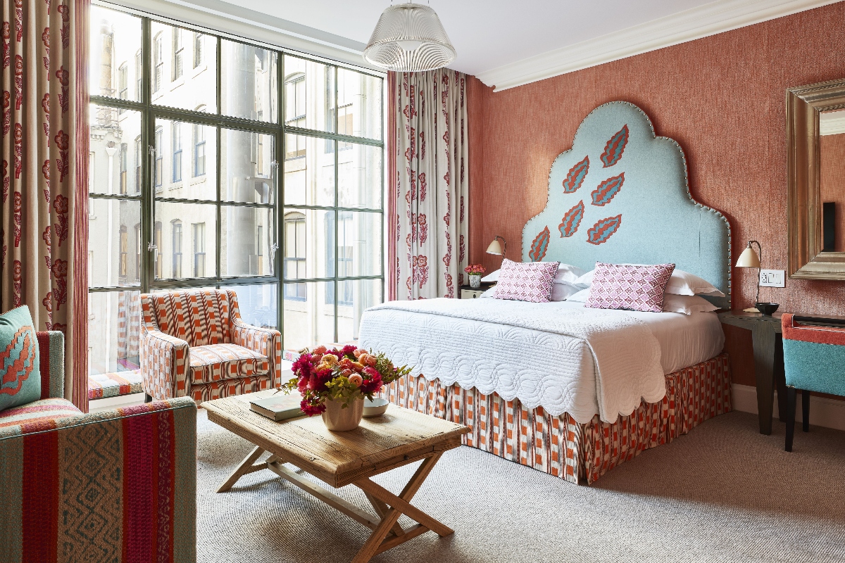 This hotel room features a distinctive curved blue headboard decorated with red paisley motifs, positioned against coral-colored walls with large multi-paned windows dressed in coordinating patterned curtains. The room includes a seating area with matching upholstered furniture in geometric patterns, a wooden bench at the foot of the bed displaying fresh flowers, and classical architectural details including crown molding and a decorative ceiling fixture.