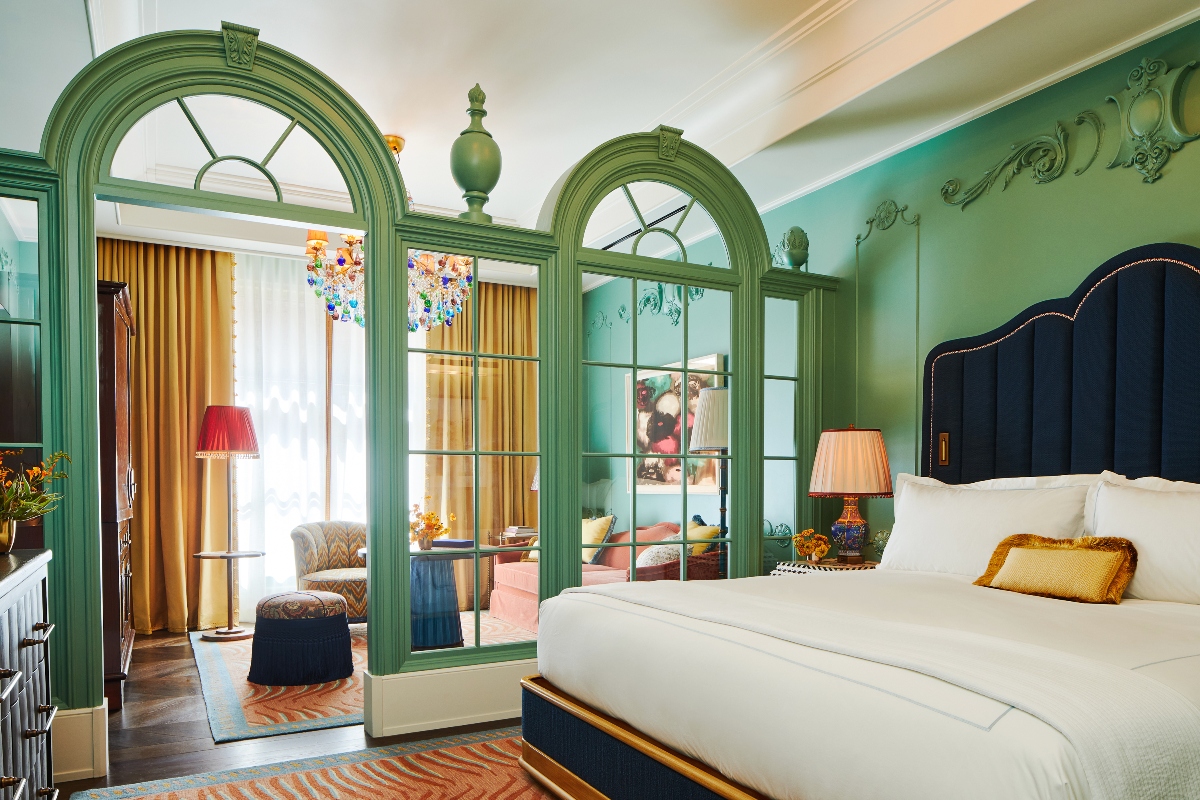 This elegantly appointed room at Fifth Avenue Hotel, one of the top luxury hotels in New York City, features distinctive jade green architectural elements including ornate arched doorways and wall panels, with a navy blue upholstered headboard and white bedding creating a striking color contrast. The space opens through decorative green-framed archways to an adjacent sitting area with golden curtains, while classical ceiling moldings and dark hardwood floors with patterned rugs complete the sophisticated interior design.