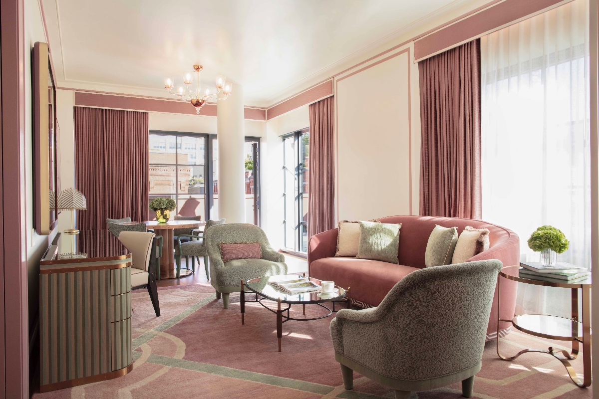This suite at Fouquet's New York, one of the newest luxury hotels in New York City, features a curved pink sofa paired with two gray textured armchairs arranged around a glass coffee table, all set on a decorative area rug with pink and green patterns. The room is illuminated by a crystal chandelier and natural light from large windows dressed in coordinating pink curtains, with French doors opening to an outdoor terrace and a dining area visible in the background.