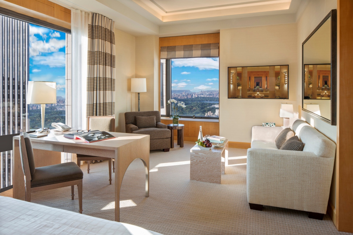 This modern hotel suite at the Four Seasons New York, one of the best luxury hotels in New York City, features a seating area with light-colored upholstered furniture, a wooden desk with chair, and large windows offering panoramic city views. The room includes floor lamps, patterned window treatments, and an adjoining bathroom visible in the background, all decorated in neutral tones with warm wood accents.