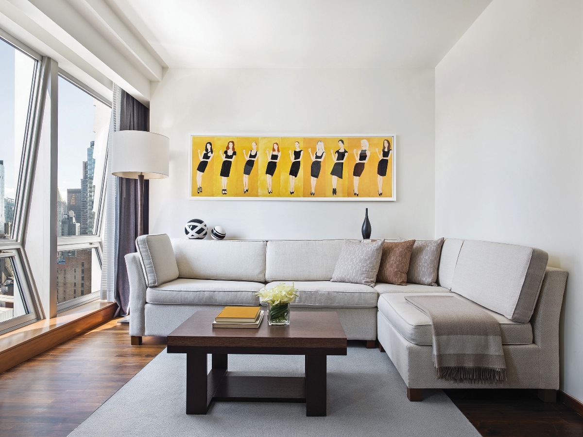 This modern hotel suite living room features a gray sectional sofa arranged around a dark wood coffee table, with floor-to-ceiling windows offering panoramic city views and dark hardwood floors throughout the space. The room is decorated with a vibrant yellow artwork depicting multiple figures in black dresses, along with contemporary furnishings including a white floor lamp and decorative accessories on the coffee table.