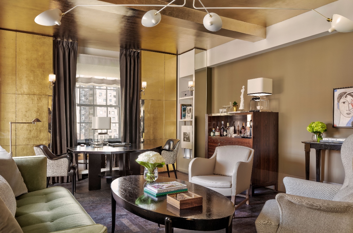 This interior of The Carlyle, one of the top luxury hotels in New York City, features a sophisticated living space with gold-toned wall panels, dark curtains framing large windows that reveal a city view, and a seating arrangement centered around a dark round coffee table. The room includes a mix of upholstered furniture in neutral tones, a dark wood bar cabinet, pendant lighting fixtures, and decorative elements including fresh white flowers and framed artwork on the walls.