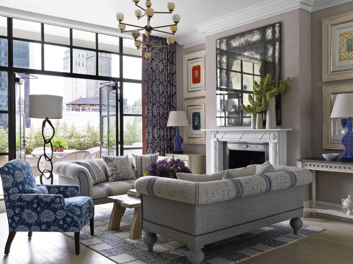This elegant hotel suite living room features a large sectional sofa in neutral tones positioned around a white fireplace, with floor-to-ceiling windows offering city views. The space includes a blue floral patterned armchair, built-in shelving with decorative objects, blue patterned curtains, and a multi-arm chandelier hanging from the ceiling.