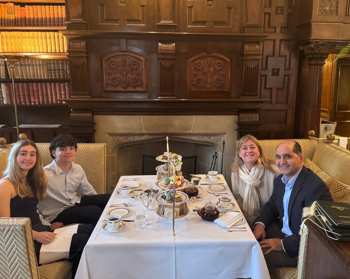 A family of four enjoys an elegant afternoon tea service at the Milestone Hotel, seated around a white-linened table featuring a traditional three-tiered serving stand filled with pastries and treats, fine china teacups, and silver service. The intimate gathering takes place in a luxurious wood-paneled library setting with floor-to-ceiling bookshelves, an ornate carved fireplace, and richly upholstered seating.