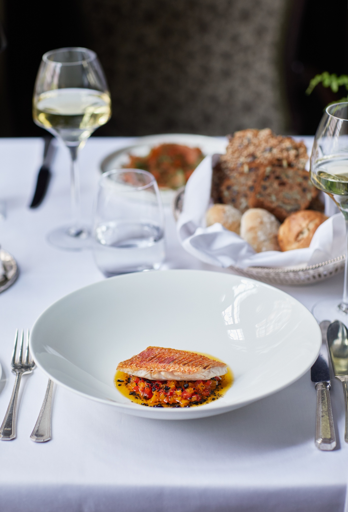 An elegantly plated fish dish with golden seared skin sits atop a colorful sauce or vegetable base in a white bowl, positioned on a formal table setting. The table features white linens, silverware, glasses of white wine, and a wicker basket containing various breads and rolls.